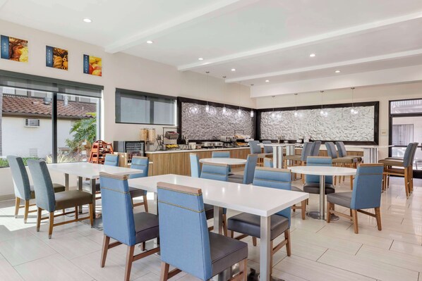 Un comedor moderno con varias mesas y sillas, una zona de cocina con mostrador, y un amplio ventanal que proporciona luz natural.