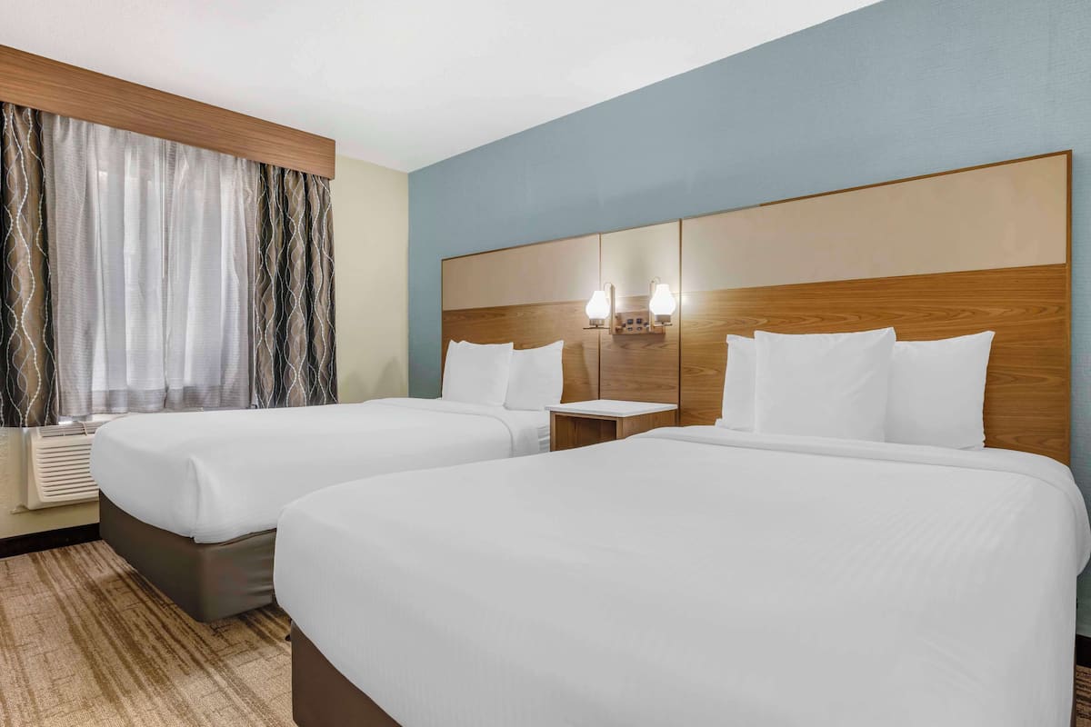 A hotel room with two beds, a wooden headboard, a nightstand with a lamp, and a window with curtains.