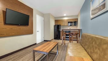 A modern kitchen with wooden cabinets, a black refrigerator, and a microwave. There is a wooden bench and a sofa in the living area.