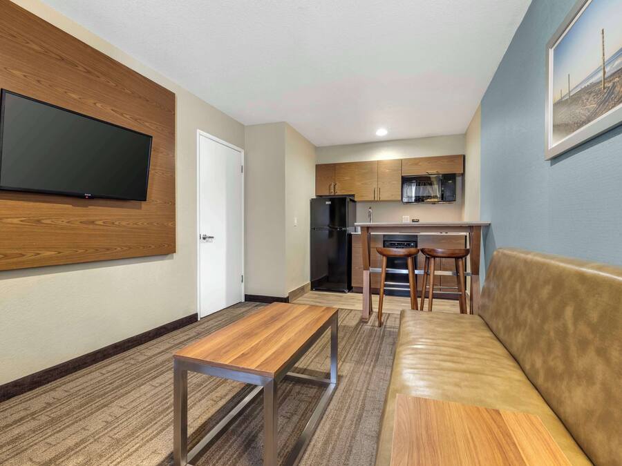 A modern kitchen with wooden cabinets, a black refrigerator, and a microwave. There is a wooden bench and a sofa in the living area.