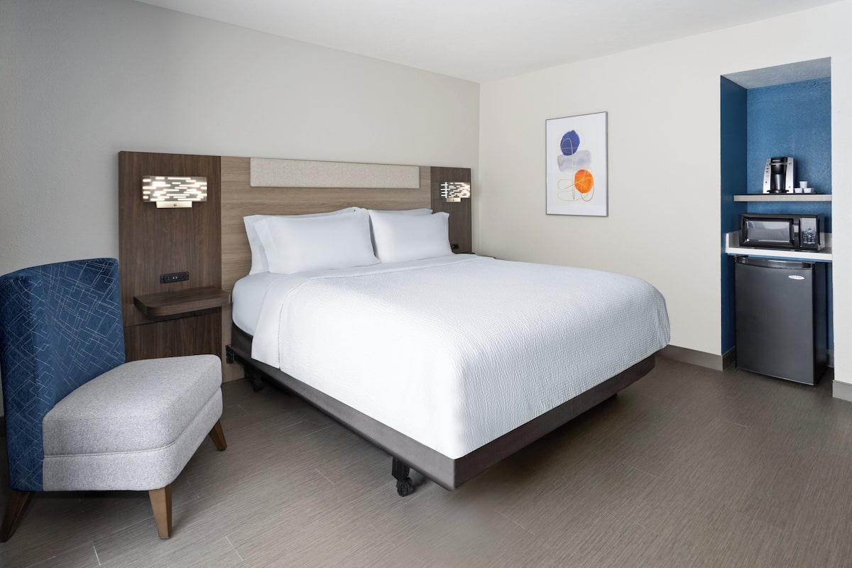 A modern hotel room with a large bed, a small desk, a chair, and a kitchenette area.
