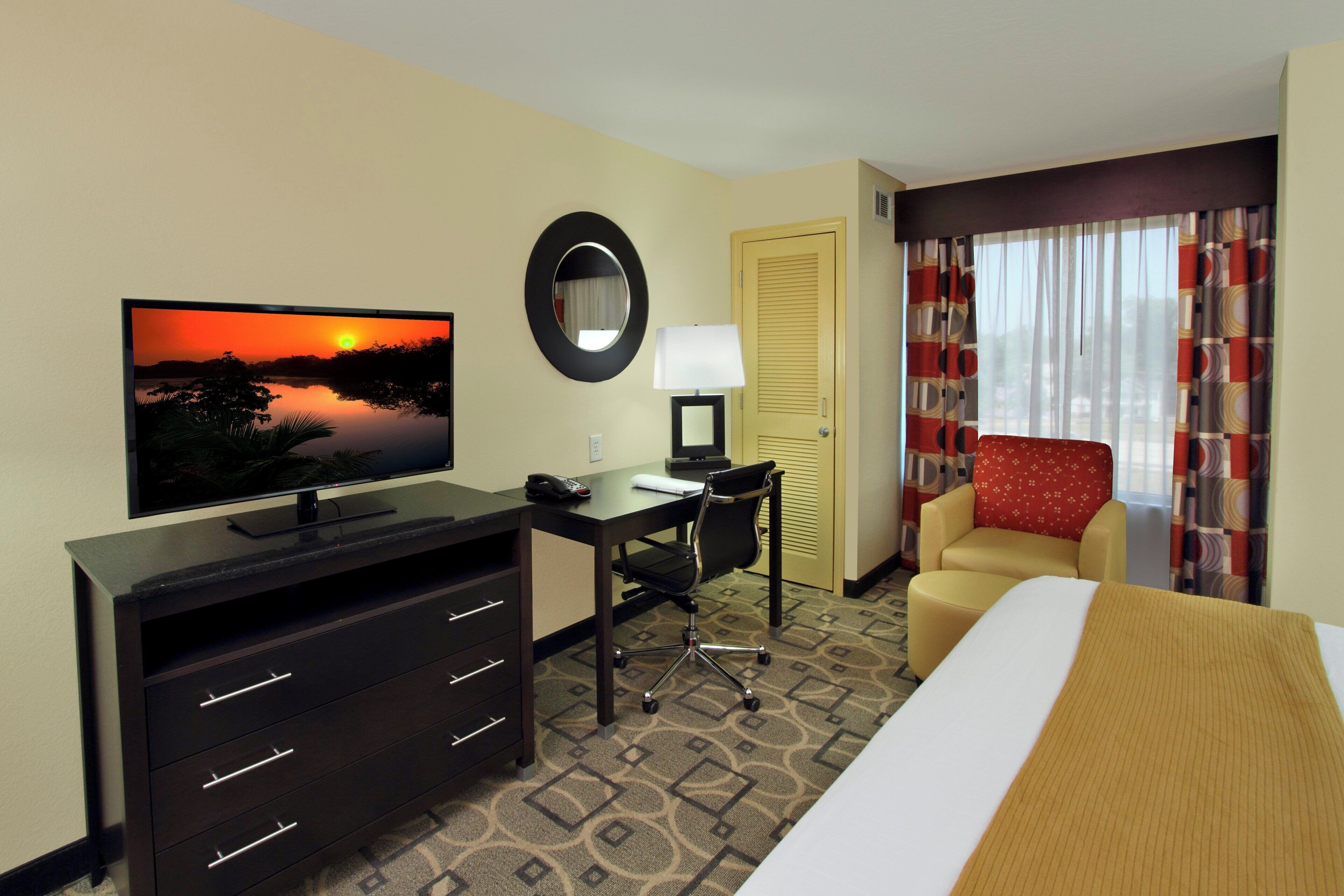 Ein Hotelzimmer mit einem Bett, einem Schreibtisch mit Stuhl, einem Fernseher, der einen Sonnenuntergang zeigt, einer Lampe und einer Tür.