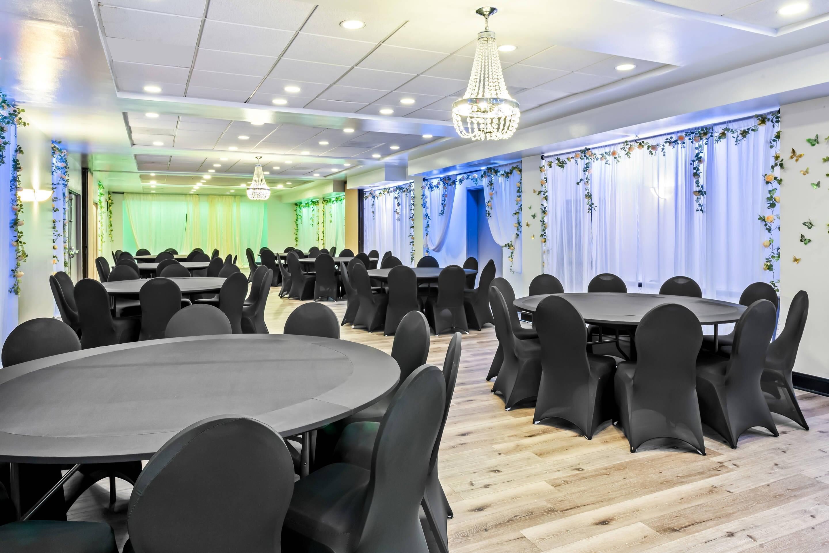 Un salón de conferencias con mesas redondas y sillas negras, una araña y cortinas con motivos florales.