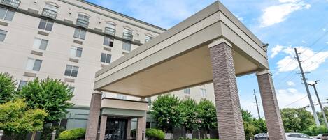 A hotel entrance with a covered walkway, a parking lot, and a building with multiple windows and balconies.