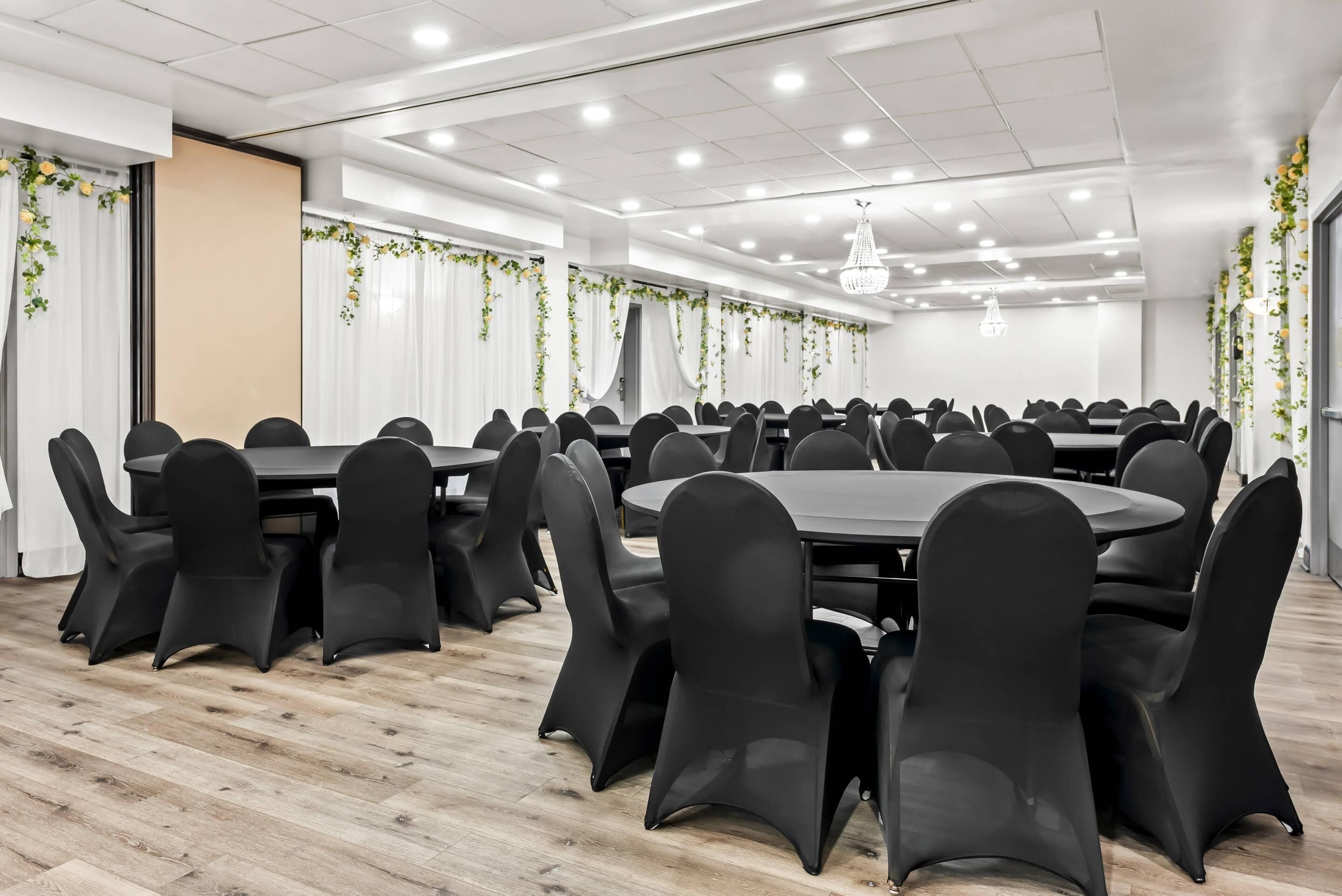 Un salón de conferencias con mesas redondas y sillas negras, cortinas blancas y plantas colgantes.