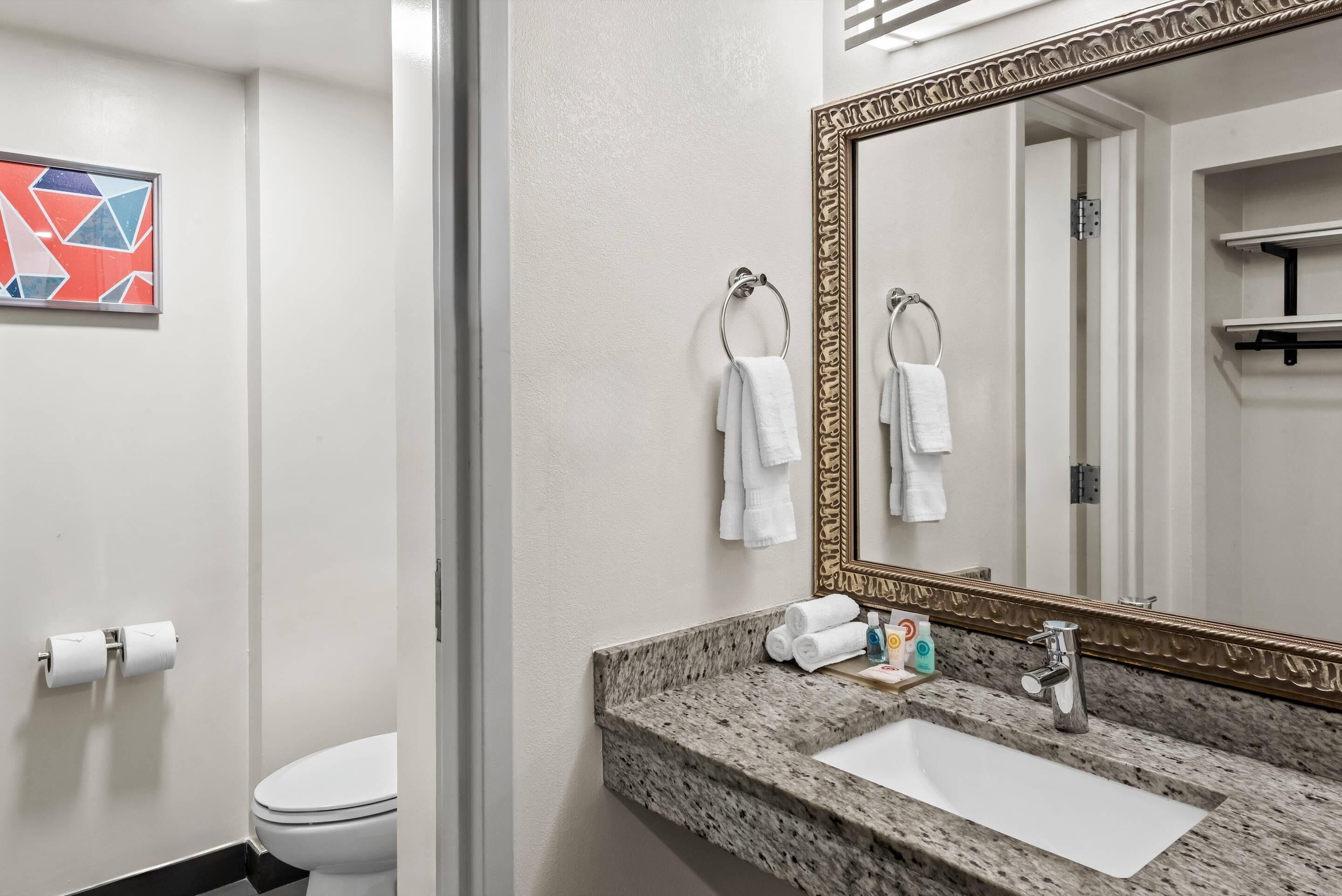 Un baño con un lavamanos blanco, un espejo grande y dos toallas colgadas en argollas.
