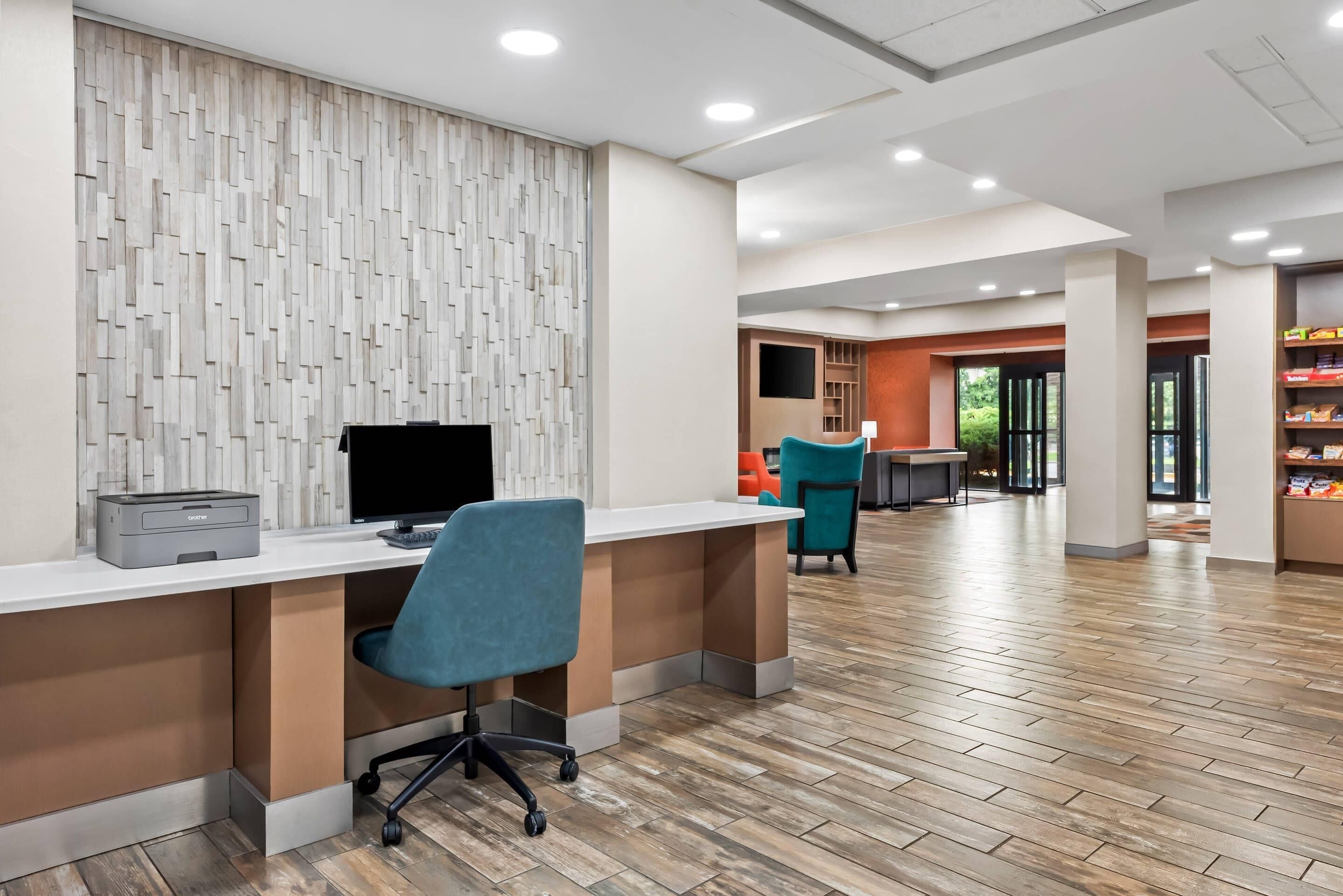 A modern office space with a reception desk, a printer, and a computer. There are two chairs, a table, and a bookshelf with items on it. The room has a wooden floor and a patterned wall.