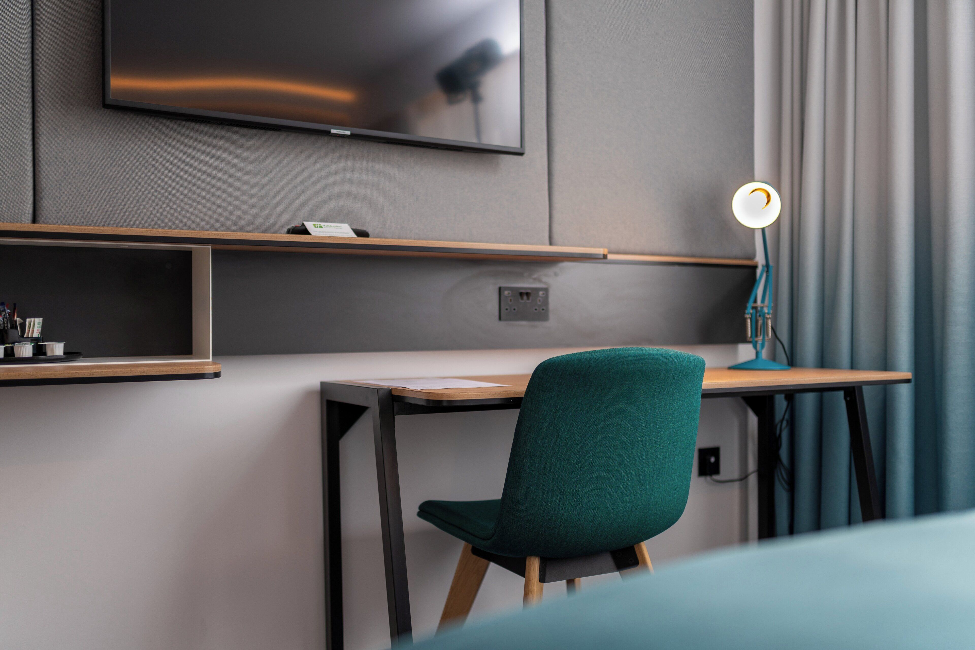 A modern hotel room with a flat-screen TV mounted on the wall, a desk with a chair, and a desk lamp.