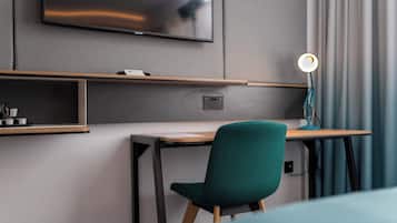 A modern hotel room with a flat-screen TV mounted on the wall, a desk with a chair, and a desk lamp.