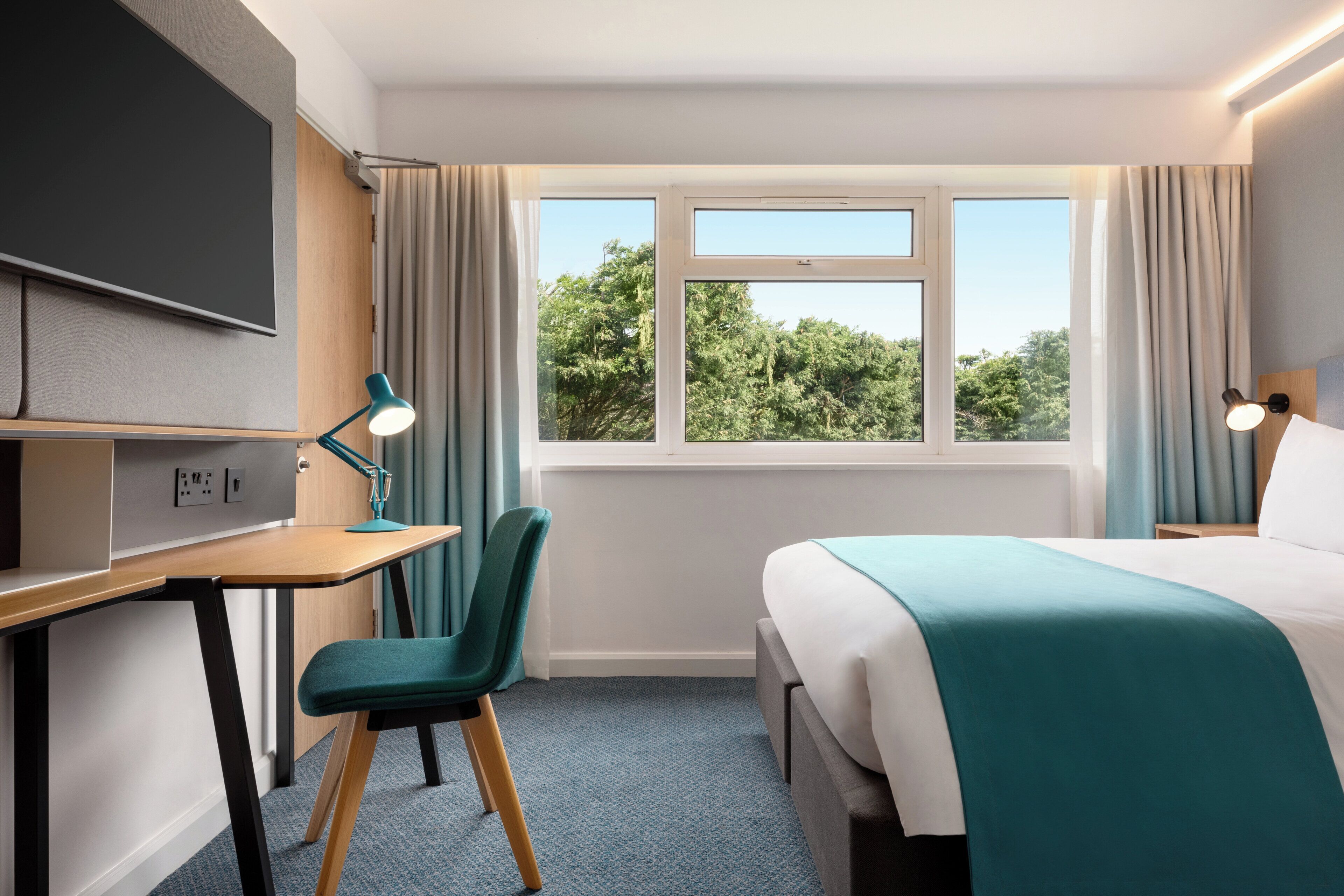 Habitación de hotel con cama, escritorio, silla y ventana con vistas a árboles.