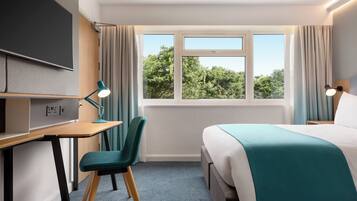 Habitación de hotel con cama, escritorio, silla y ventana con vistas a árboles.