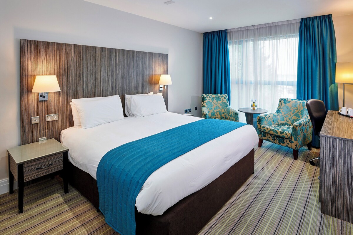 Habitación de hotel con una cama grande, dos mesitas de noche, un escritorio, una silla, una mesita pequeña con una lámpara y una ventana con cortinas.