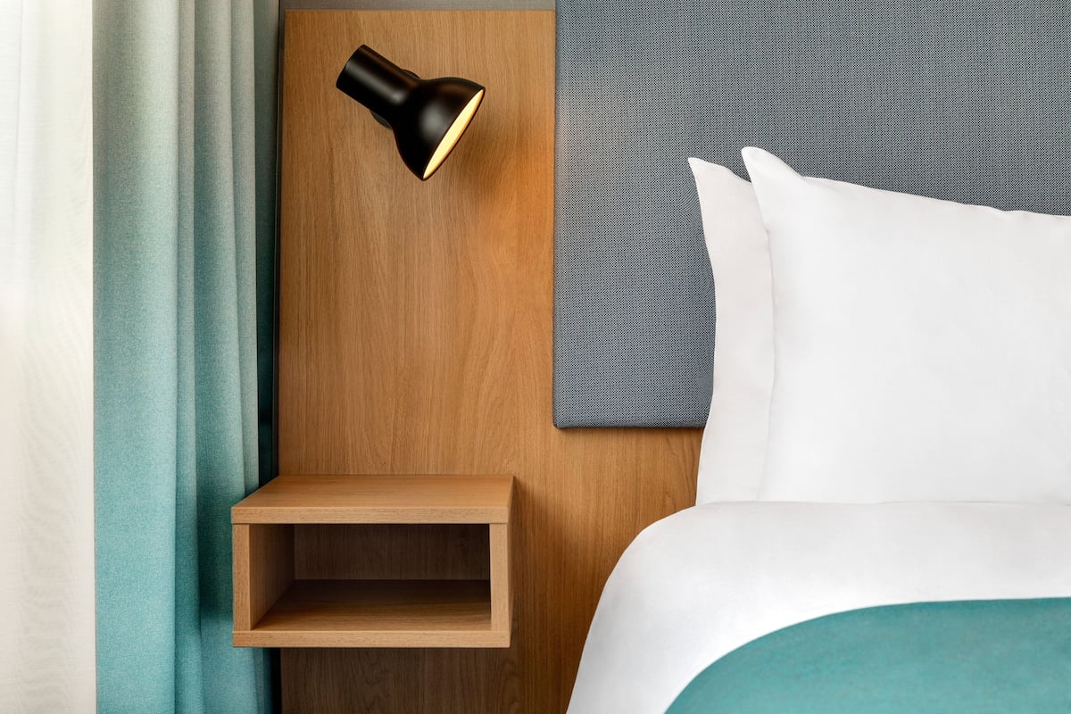 A neatly made bed with white linens, a wooden nightstand with a small shelf, a black wall-mounted lamp, and a grey headboard.