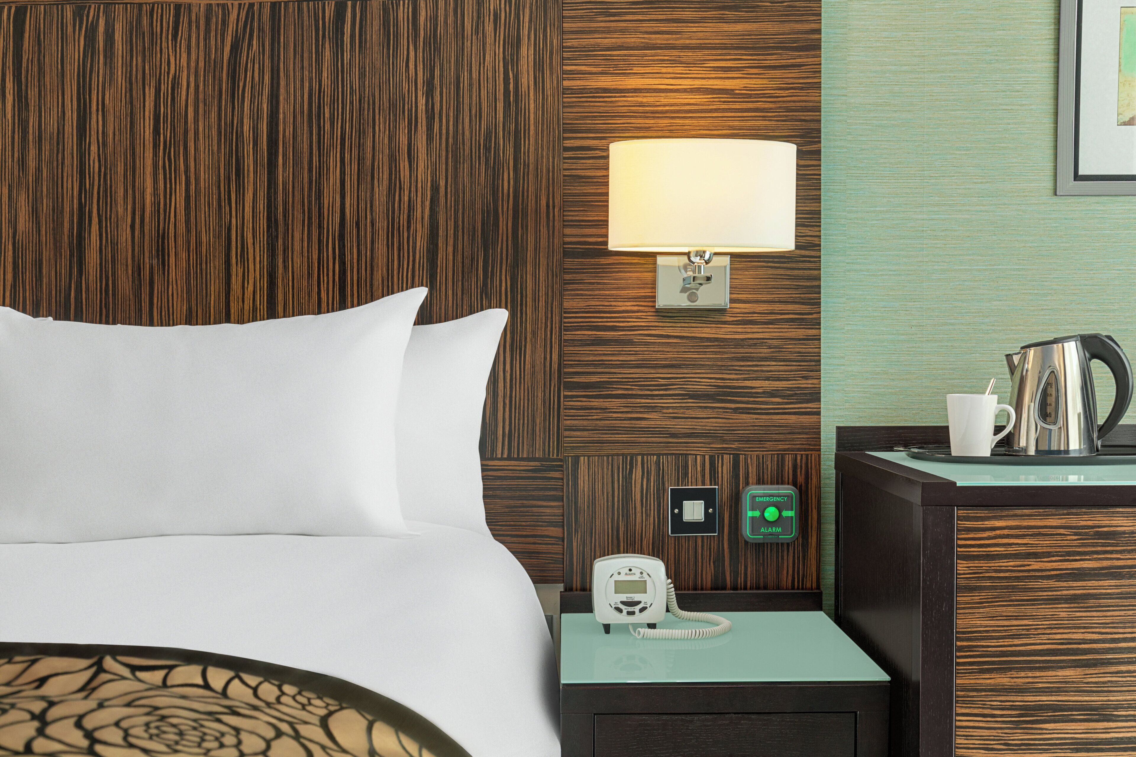 A hotel room with a bed, a nightstand with a lamp, a phone, an emergency alarm button, and a kettle.