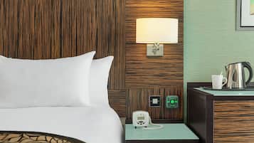 A hotel room with a bed, a nightstand with a lamp, a phone, an emergency alarm button, and a kettle.