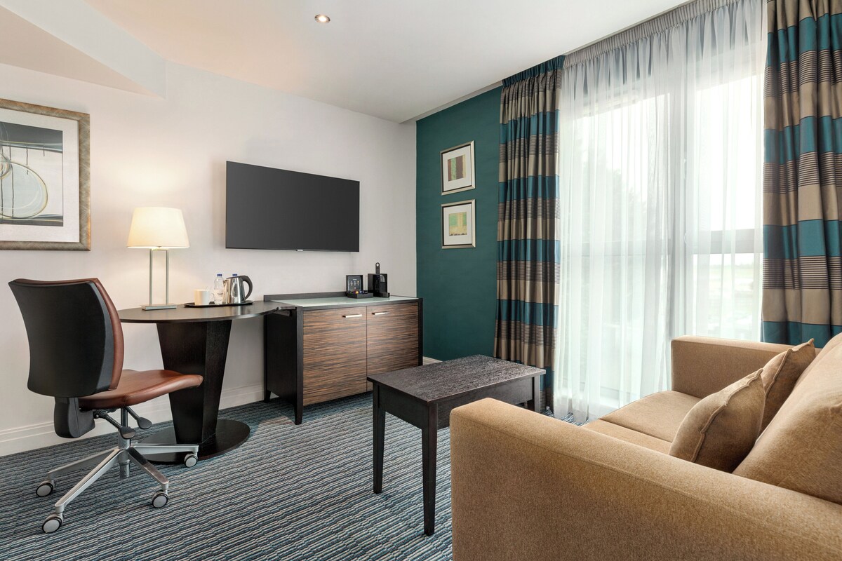 A hotel room with a desk, chair, sofa, and a flat-screen TV.