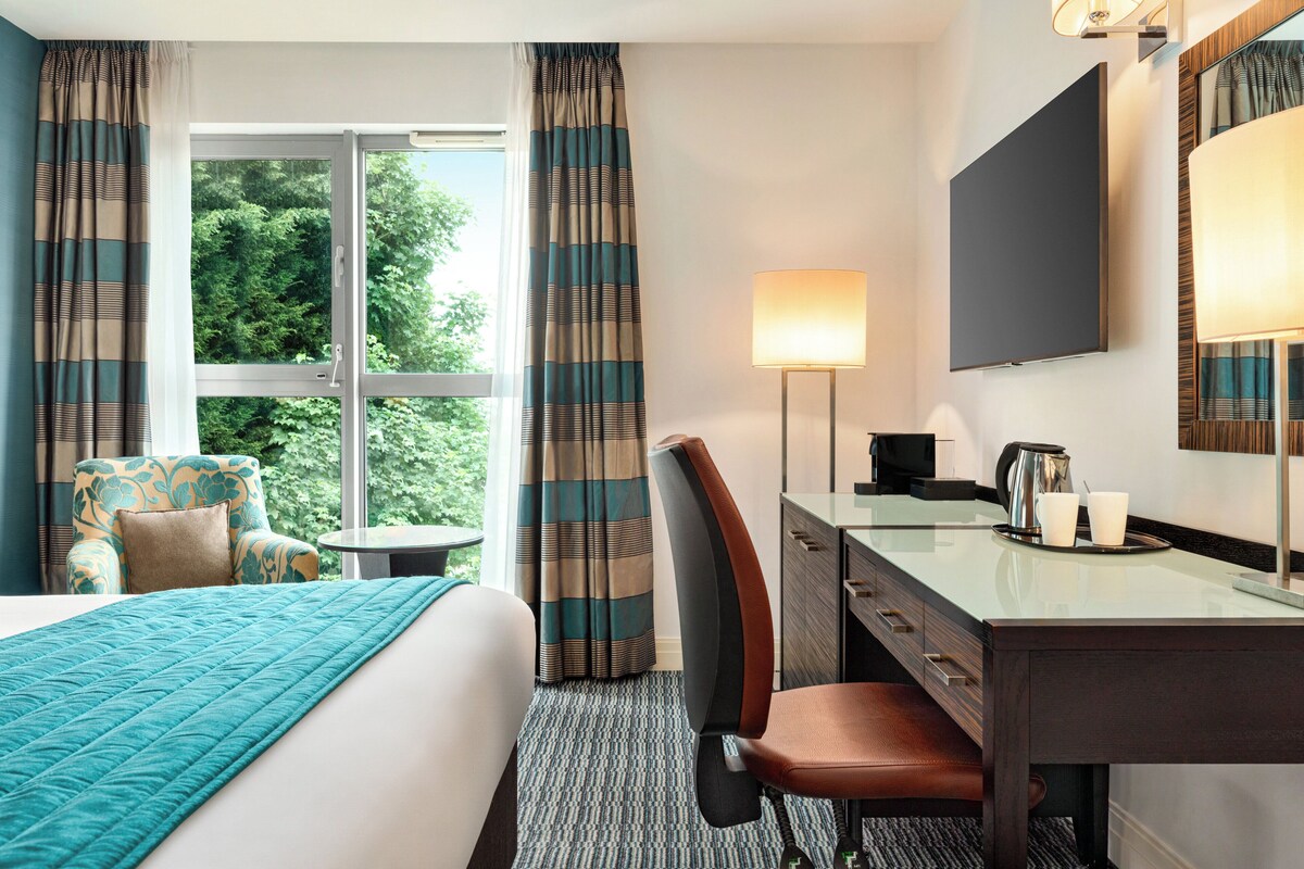 A hotel room with a bed, a desk with a lamp, a chair, and a view of greenery outside.