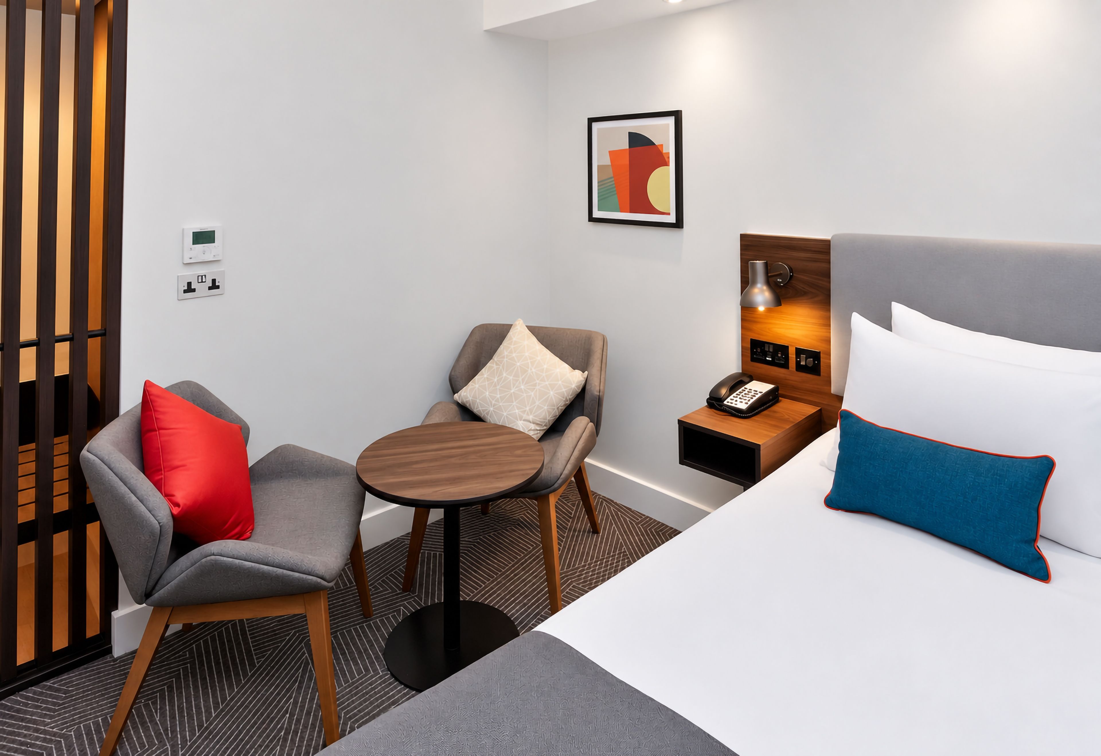 A hotel room with a bed, a desk, a chair, a small table, a nightstand, and a wall-mounted phone.