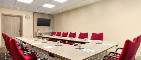 Une salle de conférence avec une longue table, des chaises rouges, une horloge et un téléviseur fixé au mur.