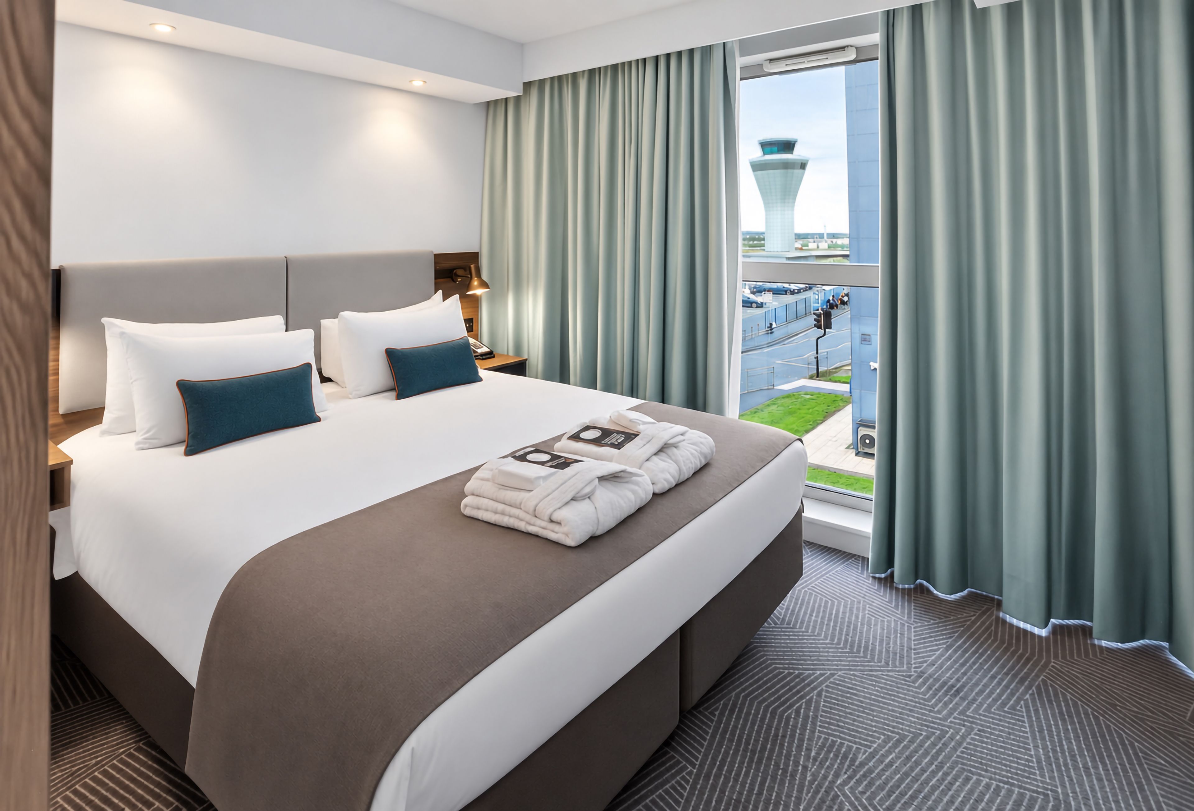 Een hotelkamer met een groot bed, een nachtkastje, een lamp en uitzicht op een stadsgezicht door het raam.