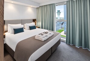 A hotel room with a large bed, a nightstand, a lamp, and a view of a cityscape through the window.