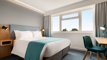 A hotel room with a bed, bedside tables, a desk with a chair, a window with curtains, and a view of greenery outside.