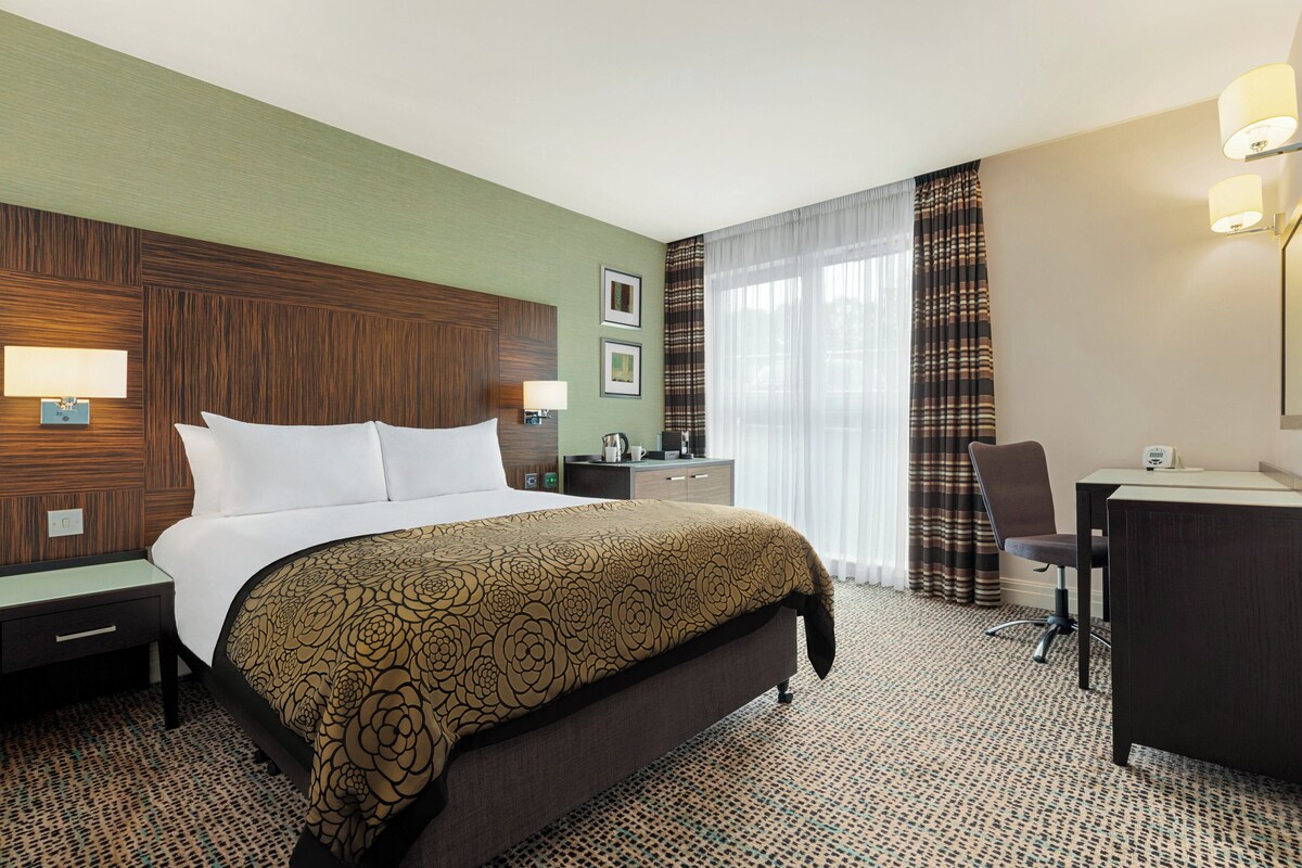 Una habitación de hotel con cama, escritorio, silla, mesita de noche, lámpara, cómoda y ventana con cortinas.