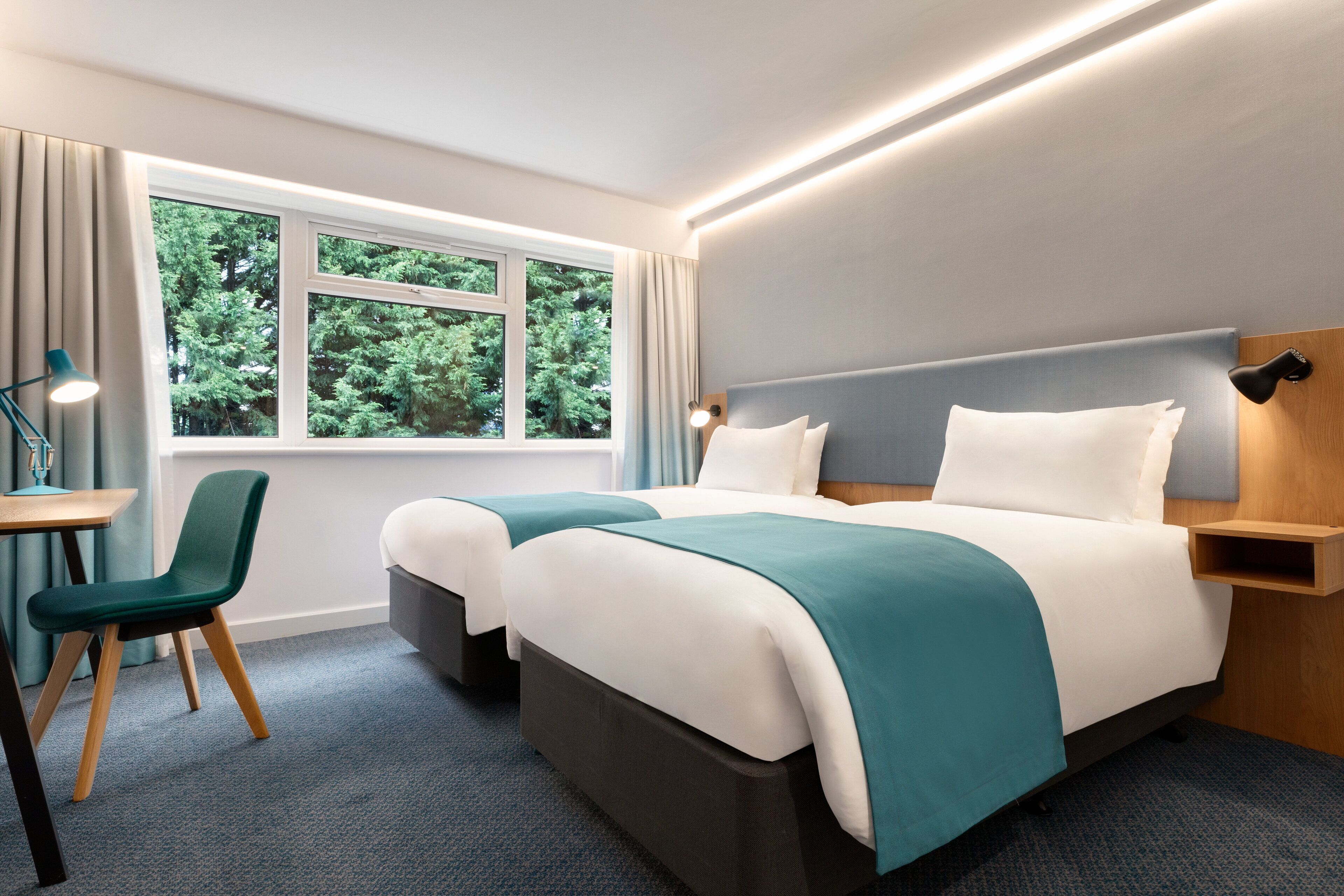 Hotellihuone, jossa on kaksi sänkyä, työpöytä, tuoli ja ikkuna, josta on näkymä puita kohti.