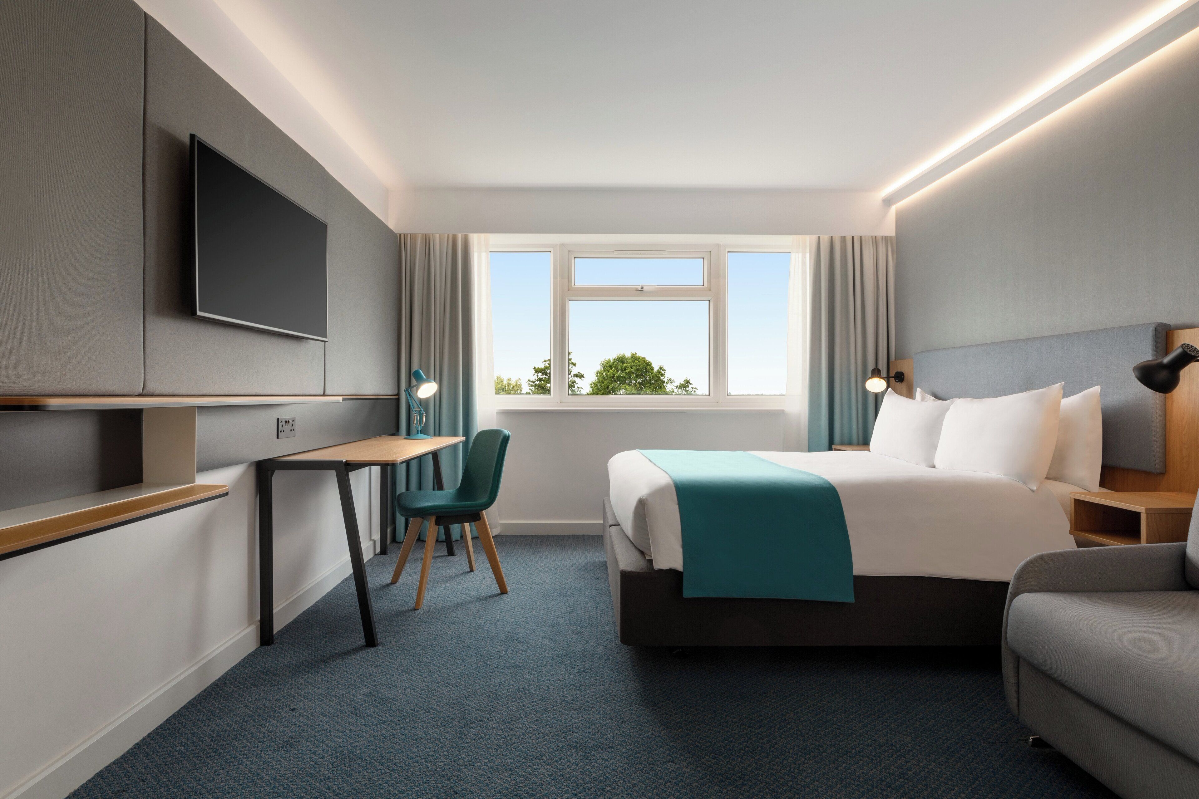 A hotel room with a bed, a desk, a chair, a TV, and a window with a view of trees.