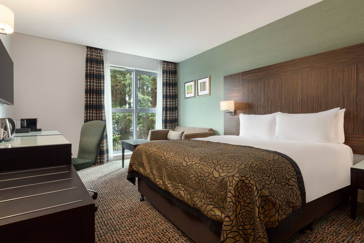 A hotel room with a large bed, a desk with a chair, a window with curtains, and a wall-mounted lamp.