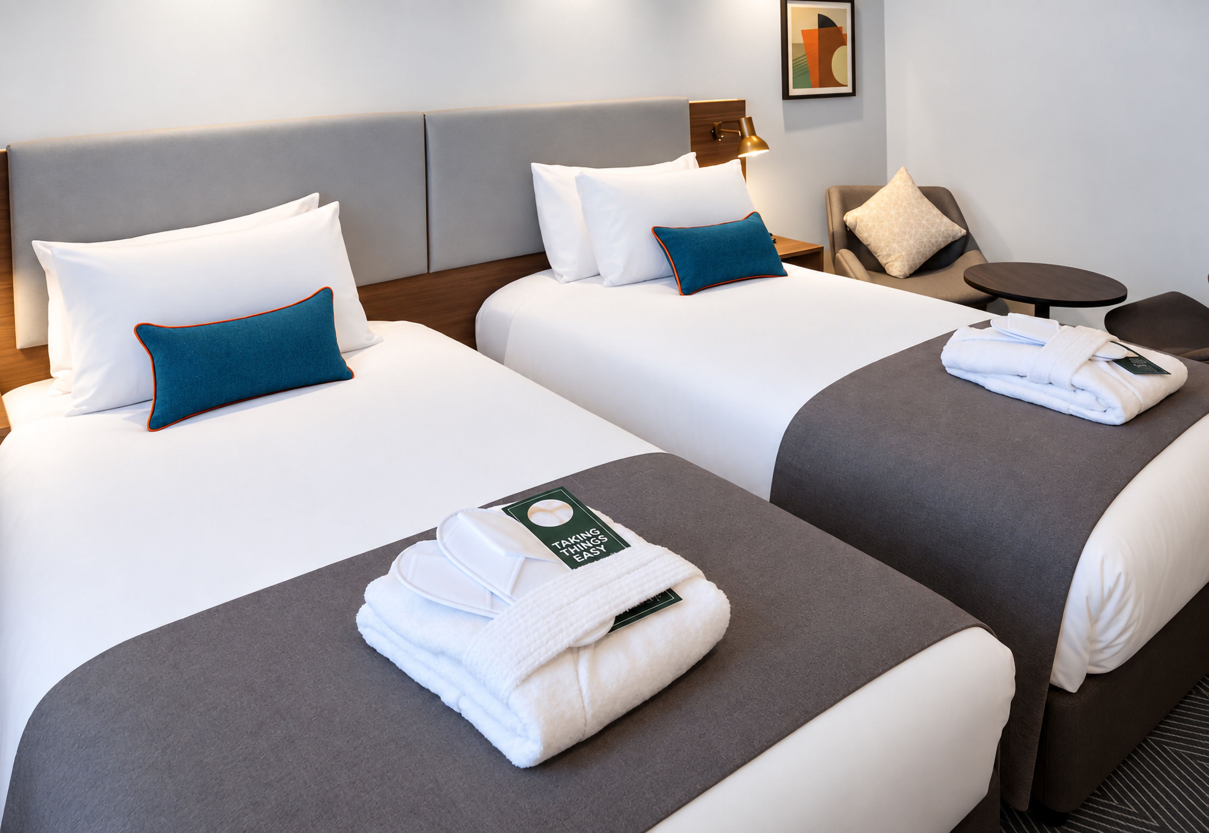 A hotel room with two beds, each with white bedding and blue pillows, a small round table, and a wall-mounted lamp.