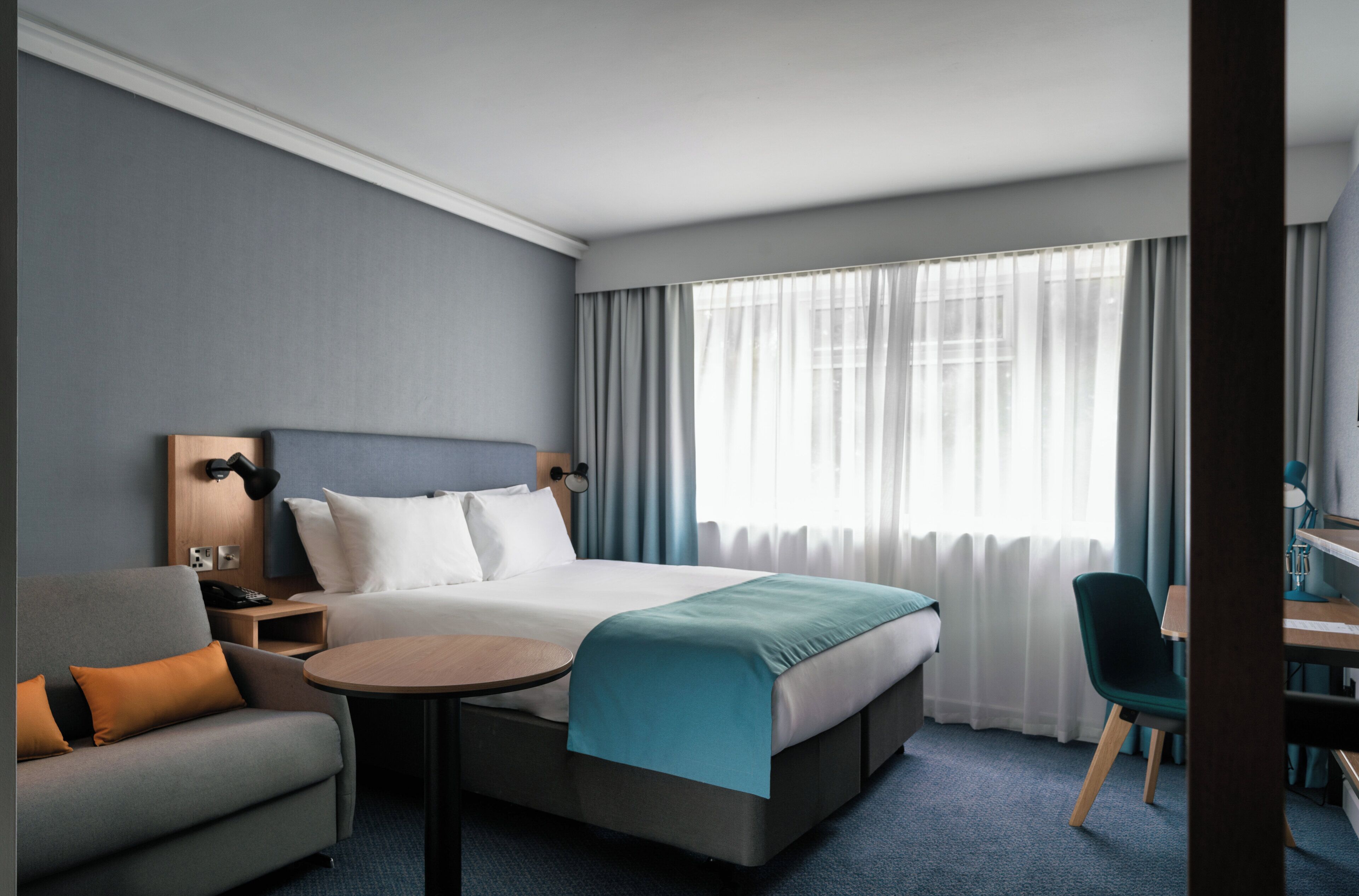 A hotel room with a bed, a desk, a chair, a small table, and a lamp.
