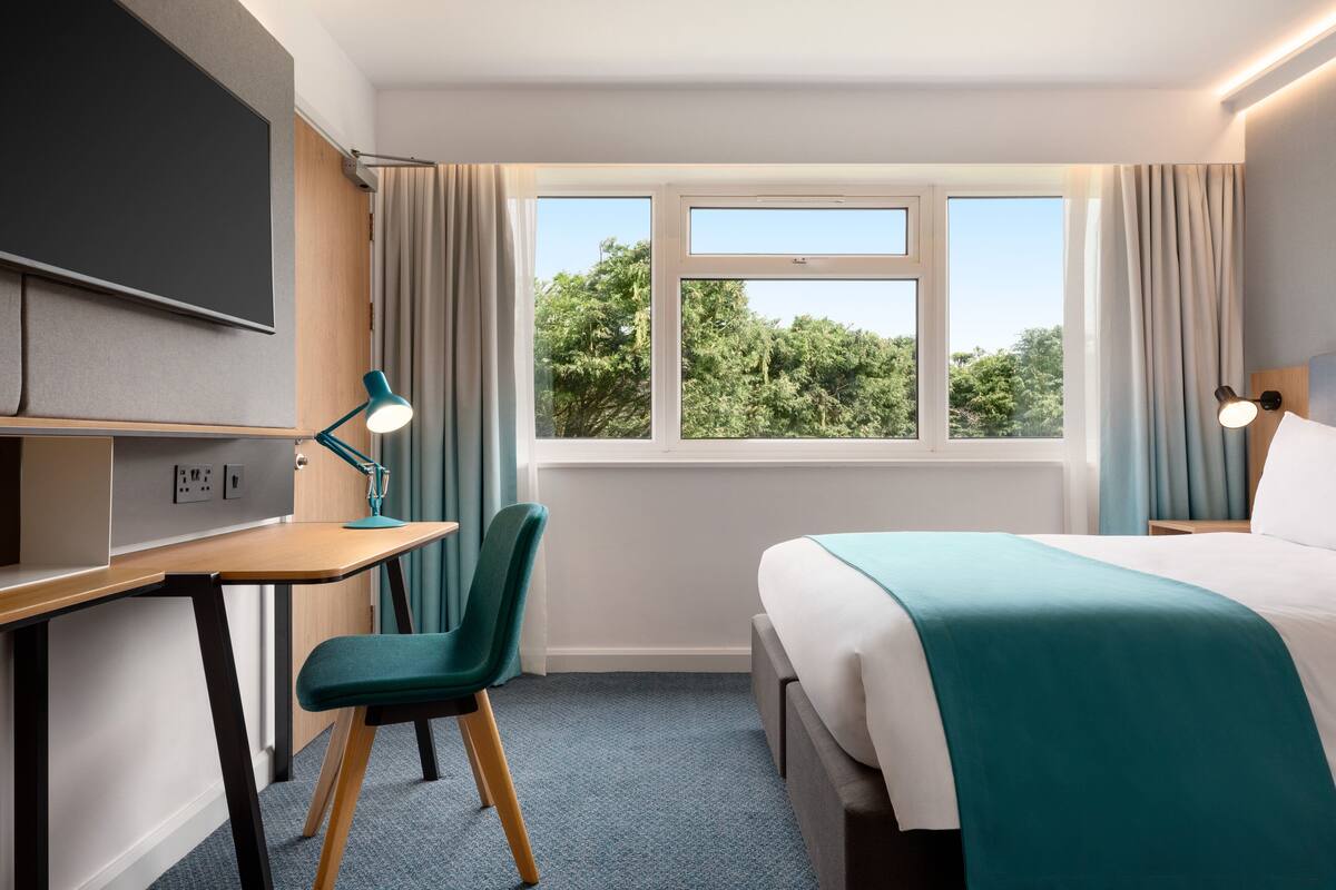 A hotel room with a bed, desk, chair, and a large window with a view of trees.