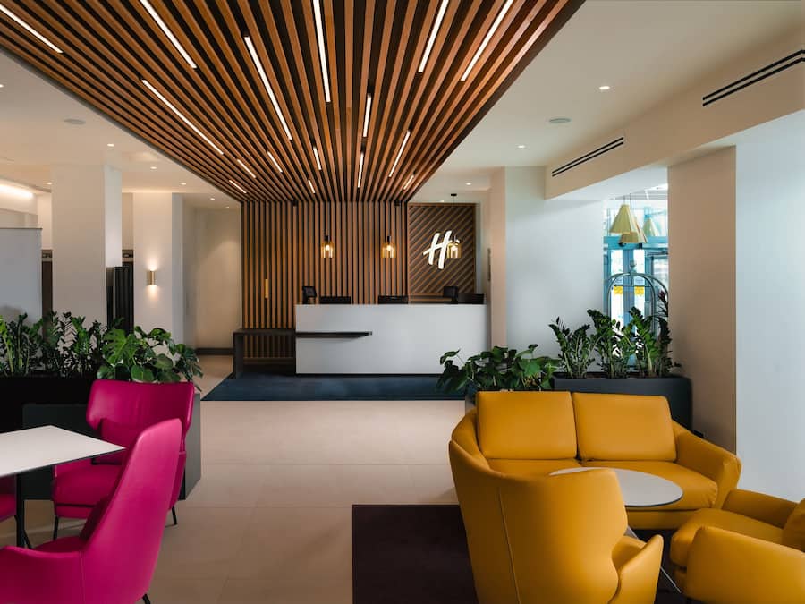 A modern reception area with a wooden slatted ceiling, a reception desk, and vibrant seating in pink and yellow.