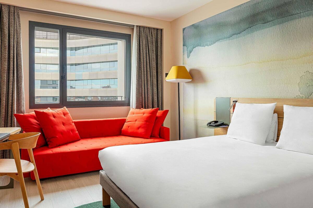 A hotel room with a bed, a red sofa, a desk, a lamp, and a large window.