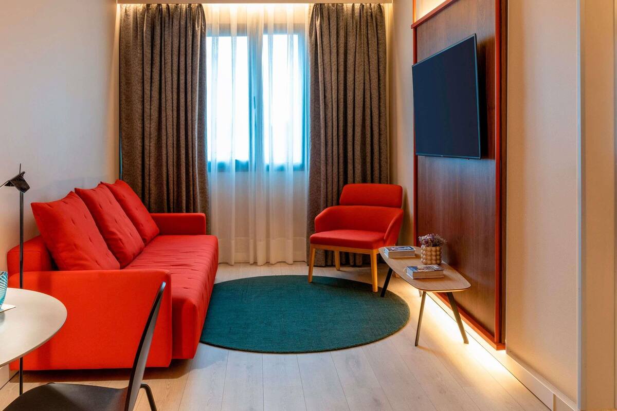 A modern living room with a red sofa, a red armchair, a small round table with books, and a TV mounted on the wall.