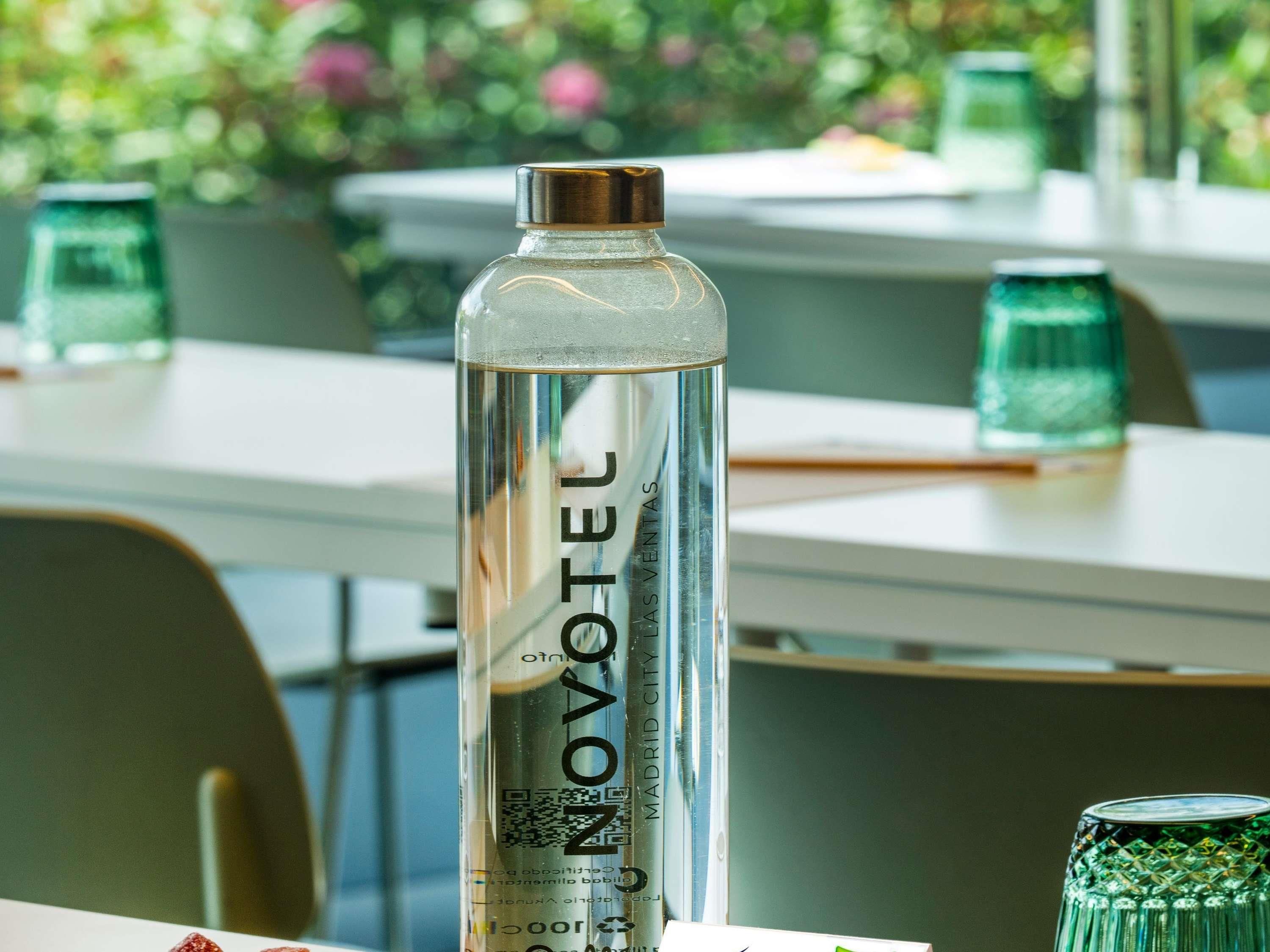 Una botella de agua transparente con tapa metálica y un código QR, sobre una mesa con un fondo borroso de un comedor.