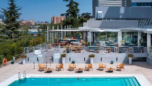 Una piscina en la azotea con sillones y mesas, rodeada de plantas en macetas y vistas panorámicas de la ciudad.