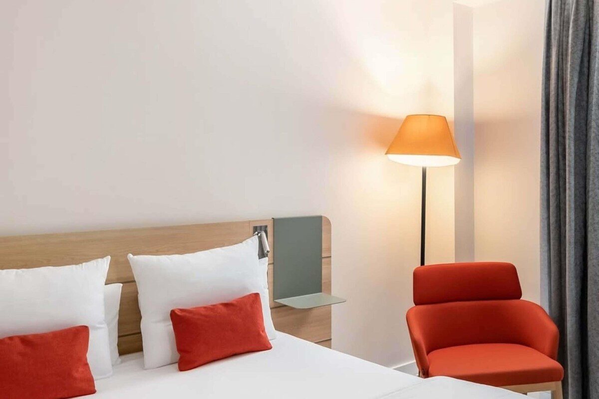 A hotel room with a bed, a nightstand, a lamp, a chair, and a curtain.