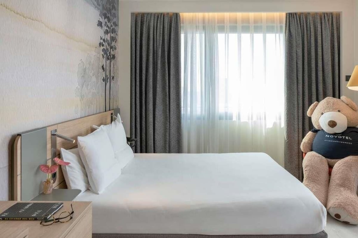 Habitación de hotel con cama, mesita de noche con lámpara, un osito de peluche y ventana con cortinas.