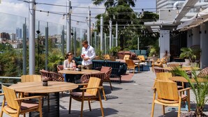 An outdoor dining area with wooden tables and chairs, surrounded by glass railings and potted plants.