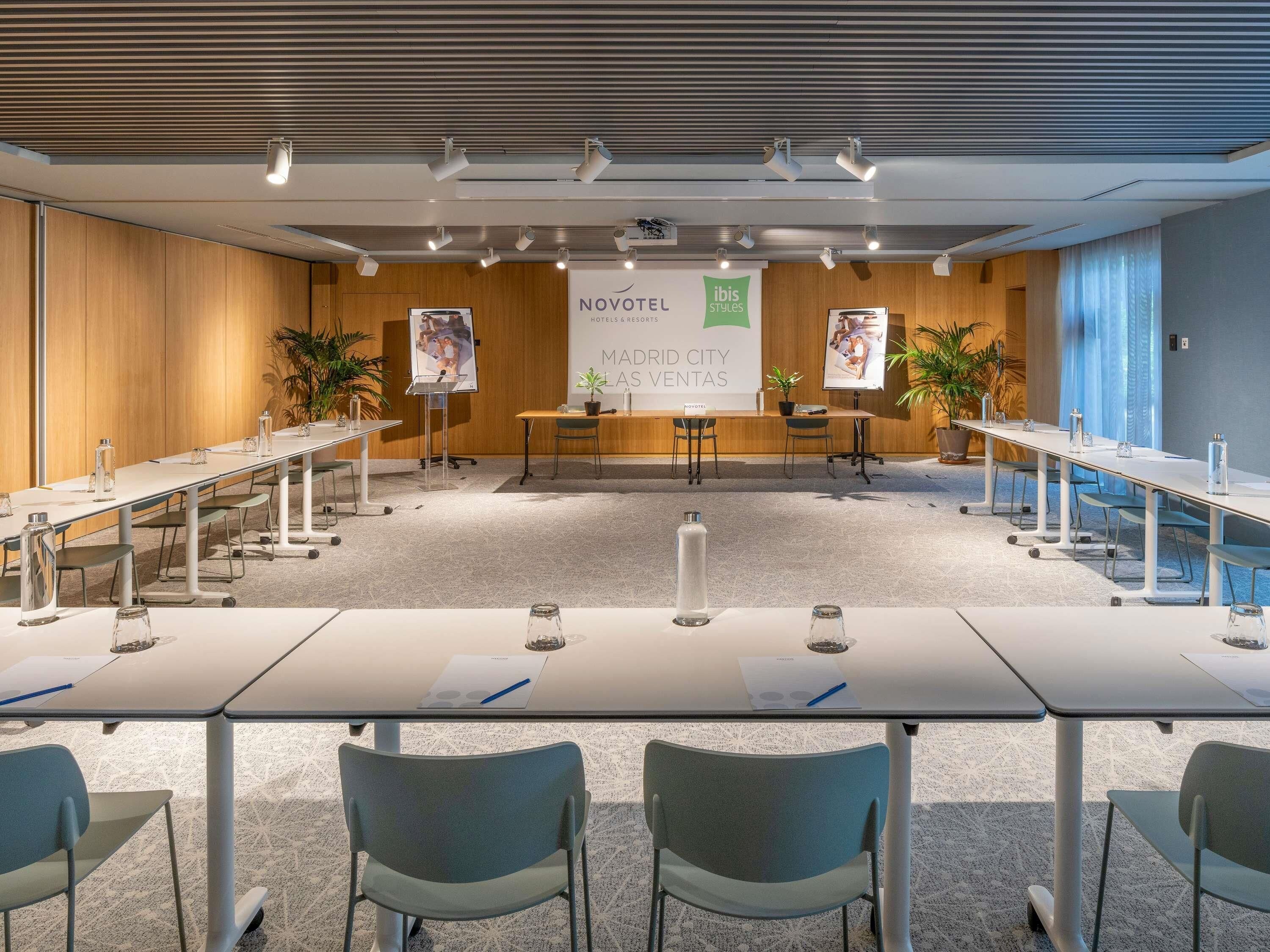 A modern conference room with long tables, chairs, and a banner advertising Novotel Madrid City Las Ventas.