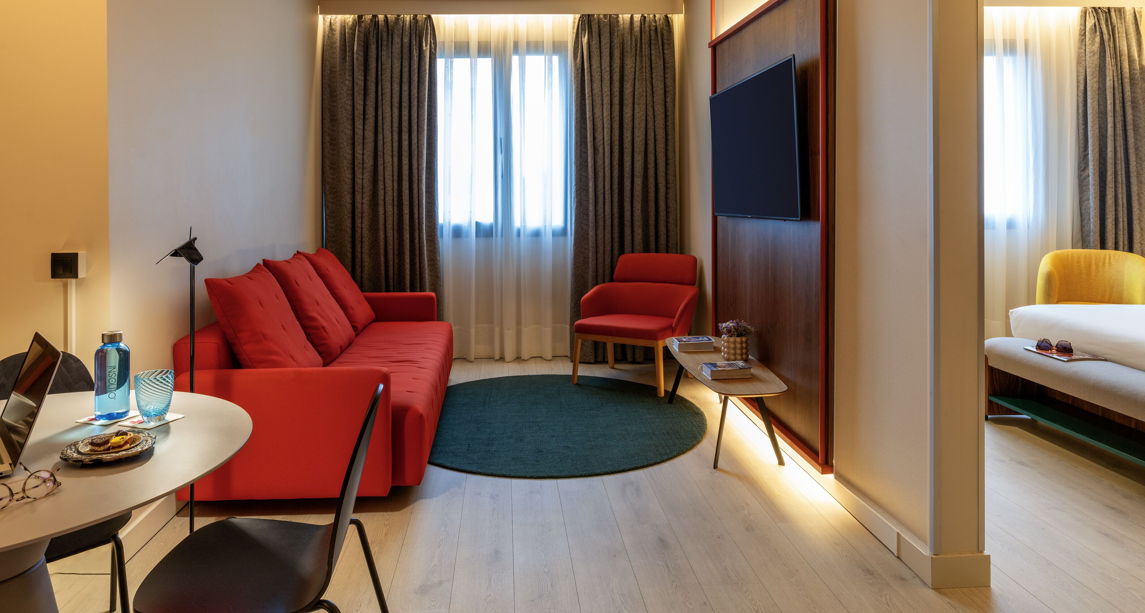 A modern hotel room with a red sofa, a small round table, a TV mounted on the wall, and a yellow armchair.