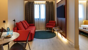 A modern hotel room with a red sofa, a small round table, a TV mounted on the wall, and a yellow armchair.