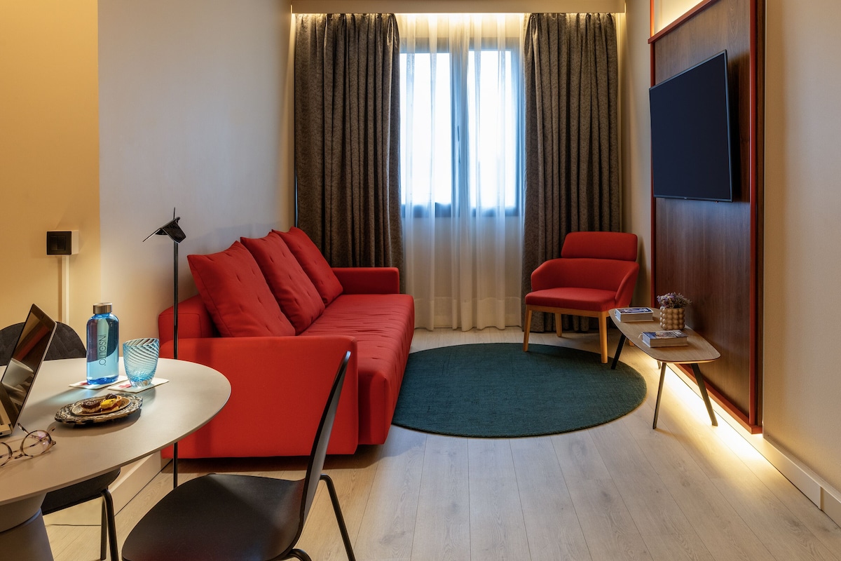 Una habitación de hotel moderna con un sofá rojo, una mesita redonda pequeña, un televisor montado en la pared y un sillón amarillo.