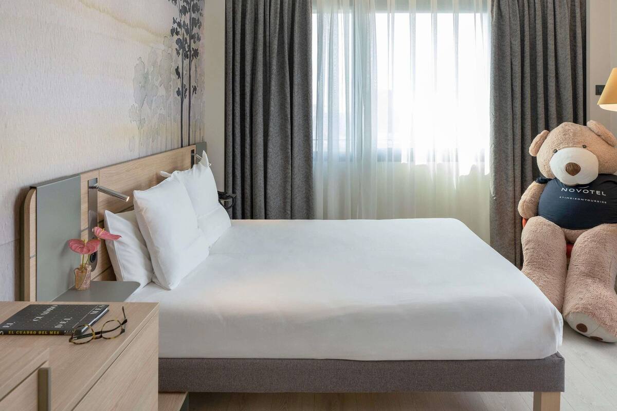 A hotel room with a bed, a bedside table with a book and glasses, a large teddy bear, and a window with curtains.