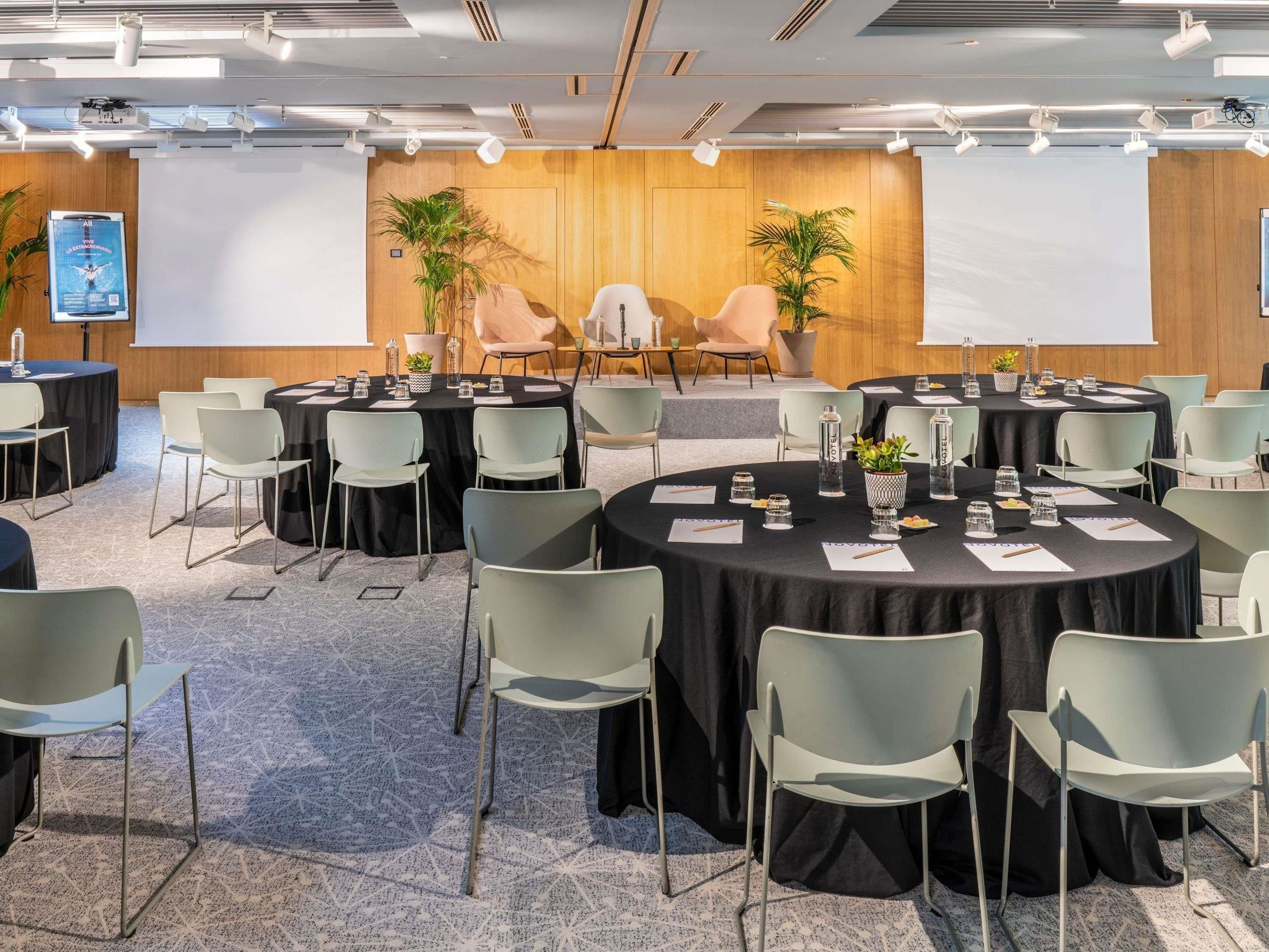 Un salón de conferencias con mesas redondas, sillas y un escenario con una pantalla.