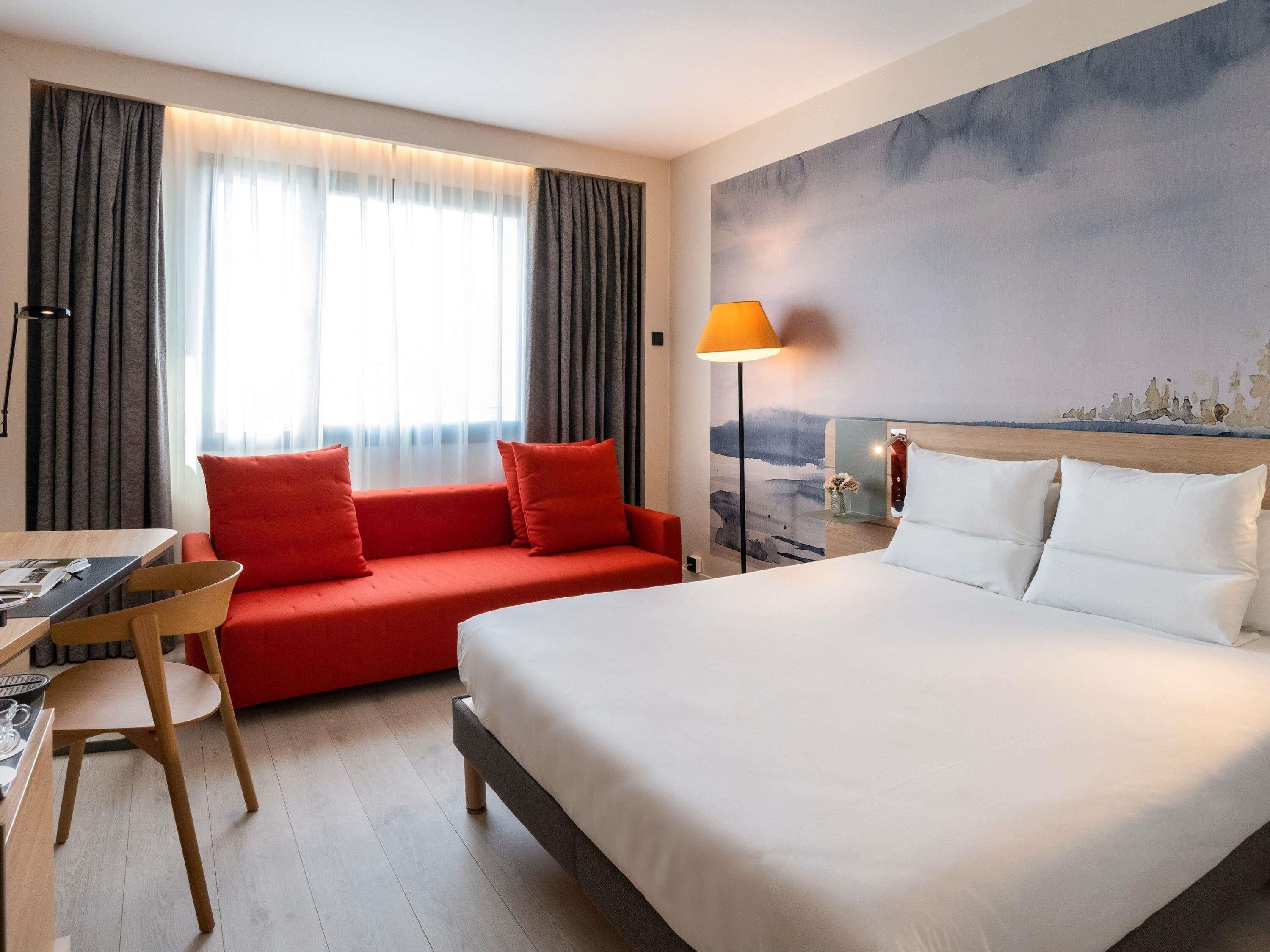A hotel room with a bed, a red sofa, a desk with a chair, a lamp, and a painting on the wall.