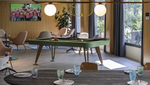 A modern room with a pool table, comfortable seating, and large windows offering a view of greenery.