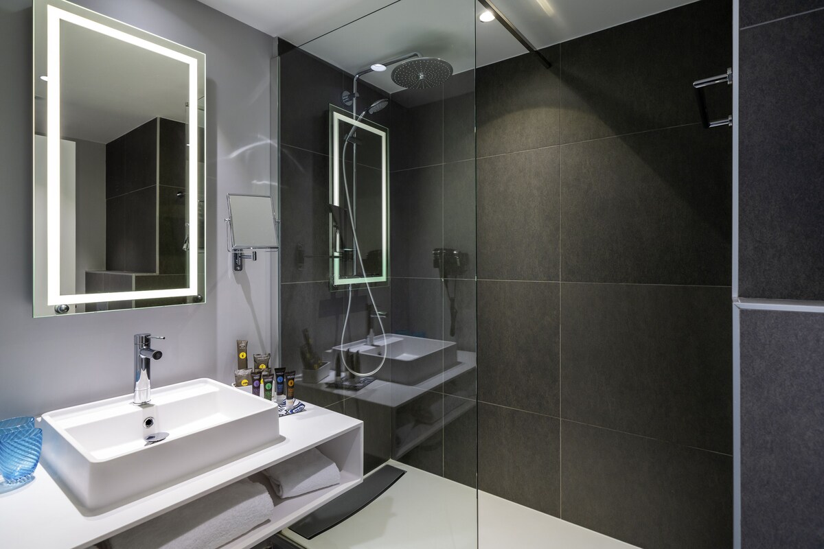 Un baño moderno con un espejo grande, dos lavamanos, una ducha y una zona de ducha con paredes de vidrio.